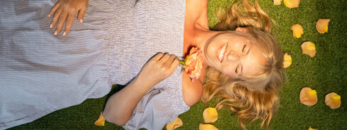 Blonde Frau liegt auf der grünen Wiese mit gestreuten Rosenblättern und lacht in die Kamera