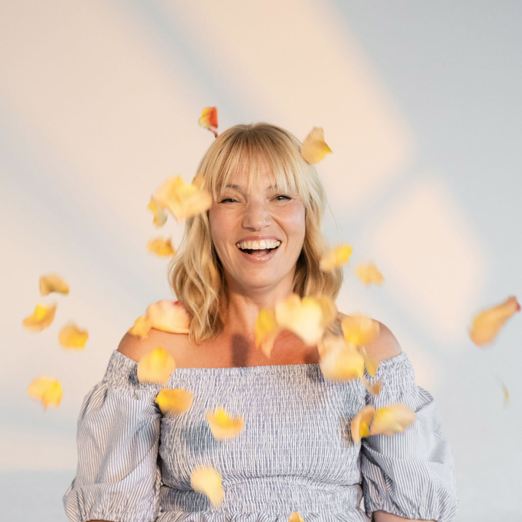 Lachende Frau wirft gelb orange farbene einzelne Rosenblätter in die Luft und sitzt auf einer grünen Wiese