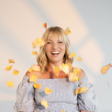 Lachende Frau wirft gelb orange farbene einzelne Rosenblätter in die Luft und sitzt auf einer grünen Wiese