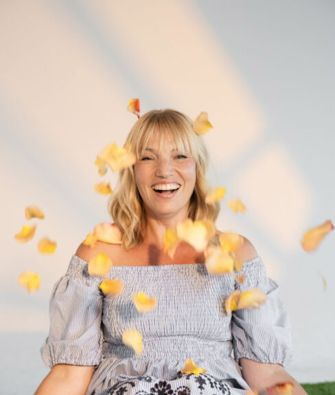 Lachende Frau wirft gelb orange farbene einzelne Rosenblätter in die Luft und sitzt auf einer grünen Wiese