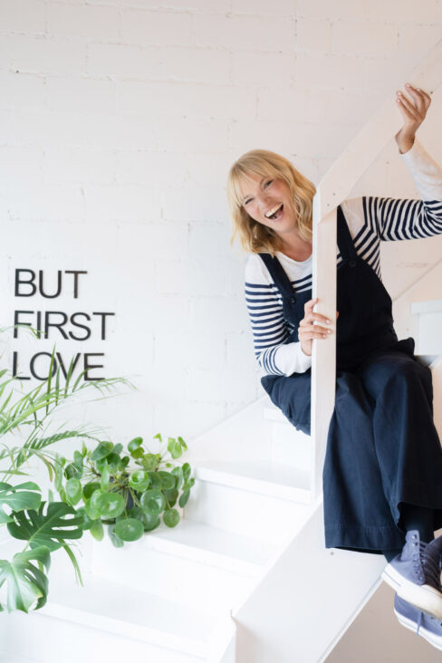 Blonde Frau in Latzhose und Ringelshirt schaut lachend über ein Treppengeländer.