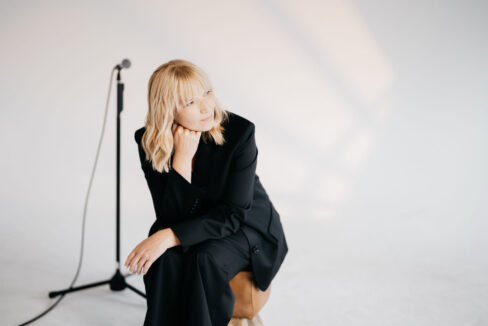 Blonde Frau im blauen Hosenanzug mit Mikrofon auf Mikrofonständer im Hintergrund.