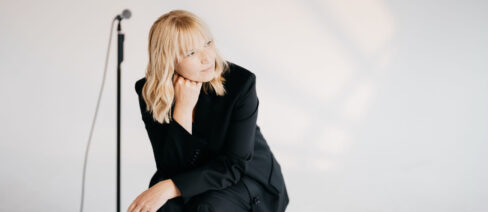 Blonde Frau im blauen Hosenanzug mit Mikrofon auf Mikrofonständer im Hintergrund.